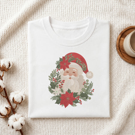 A folded white crewneck t-shirt with a festive embroidered Santa Claus design, displayed on a textured cream fabric background with decorative cotton bolls and a wooden cup.