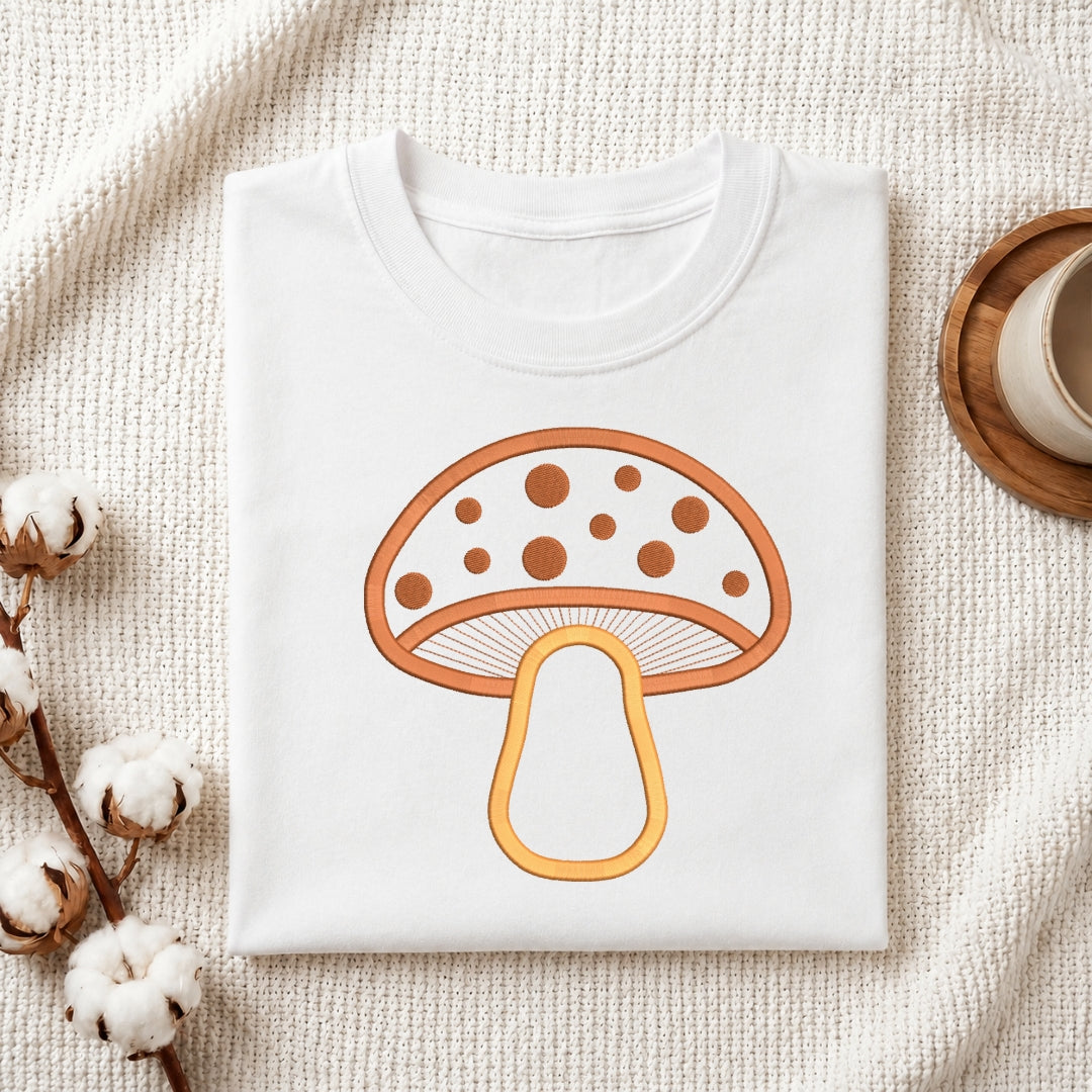 A folded white crewneck t-shirt featuring a retro-style orange and yellow mushroom graphic on the front, styled on a textured cream knit blanket with cotton bolls and a wooden cup in the background.
