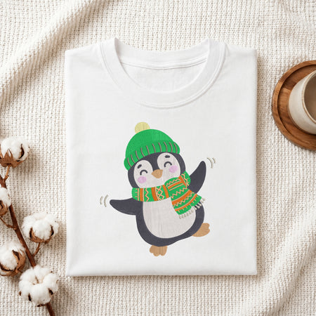 A folded white crewneck t-shirt featuring a cute cartoon penguin wearing a green knit hat and a green, orange, and white patterned scarf. The t-shirt is styled in a cozy flat-lay arrangement on a textured cream knit blanket, with cotton bolls on the left and a wooden cup on a coaster on the right.