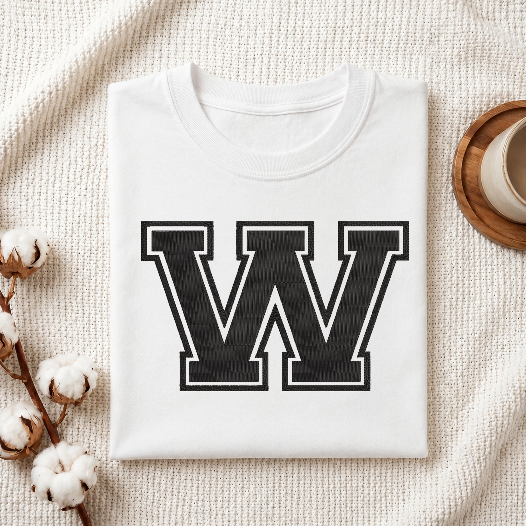 A folded white crewneck t-shirt with a large black collegiate-style letter 'W' printed on the front, styled on a textured cream knit blanket with cotton bolls and a cup in the background.