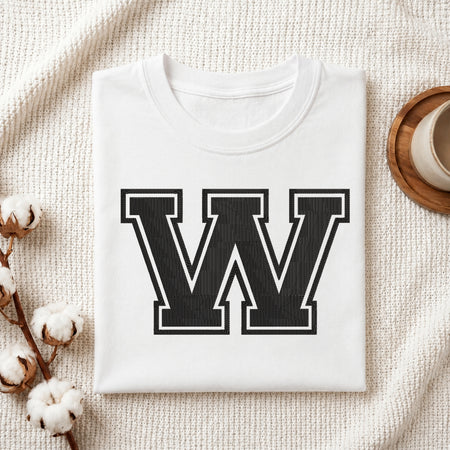 A folded white crewneck t-shirt with a large black collegiate-style letter 'W' printed on the front, styled on a textured cream knit blanket with cotton bolls and a cup in the background.