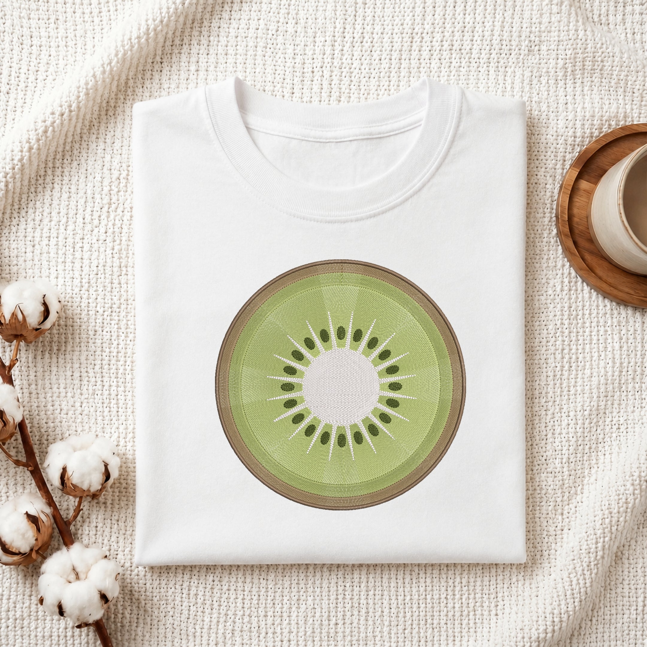 A folded white T-shirt laid flat on a textured cream fabric background, featuring a large kiwi slice graphic printed on the chest. Cotton bolls and part of a wooden cup are visible as styled props around the shirt.