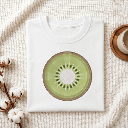 A folded white T-shirt laid flat on a textured cream fabric background, featuring a large kiwi slice graphic printed on the chest. Cotton bolls and part of a wooden cup are visible as styled props around the shirt.