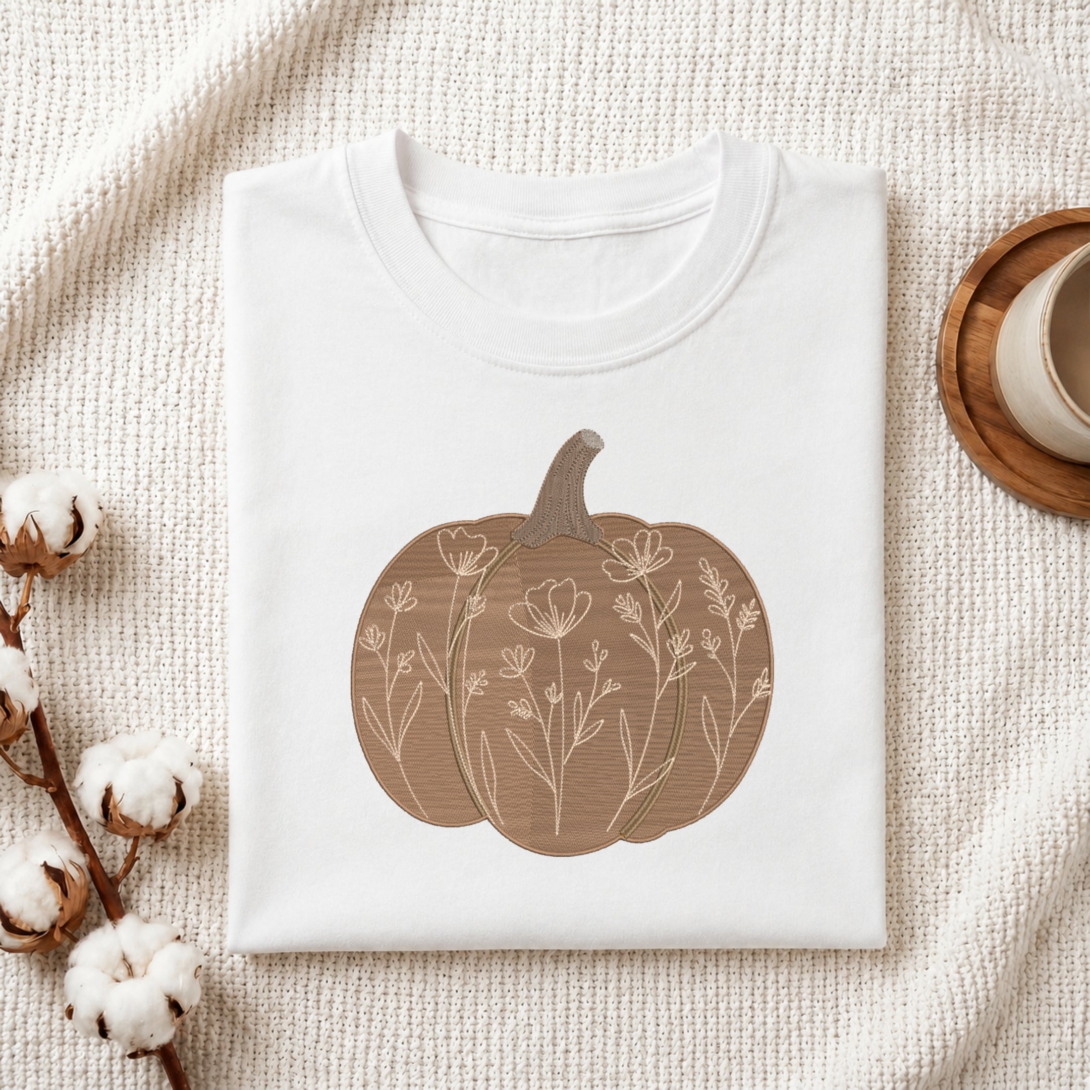 A folded white crewneck t-shirt with a brown pumpkin graphic containing floral line art, styled on a textured cream blanket with cotton bolls and a wooden cup in the background.