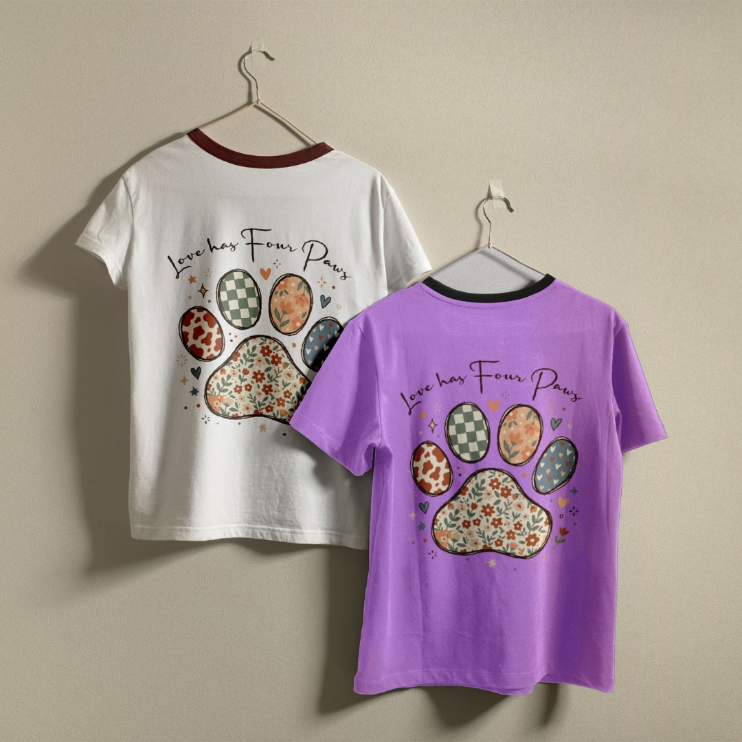 Two children's short-sleeve ringer T-shirts hanging on hangers against a plain beige wall. The left shirt is white with a dark red collar trim and features a colorful paw print and the text 'Love has Four Paws' on the front.