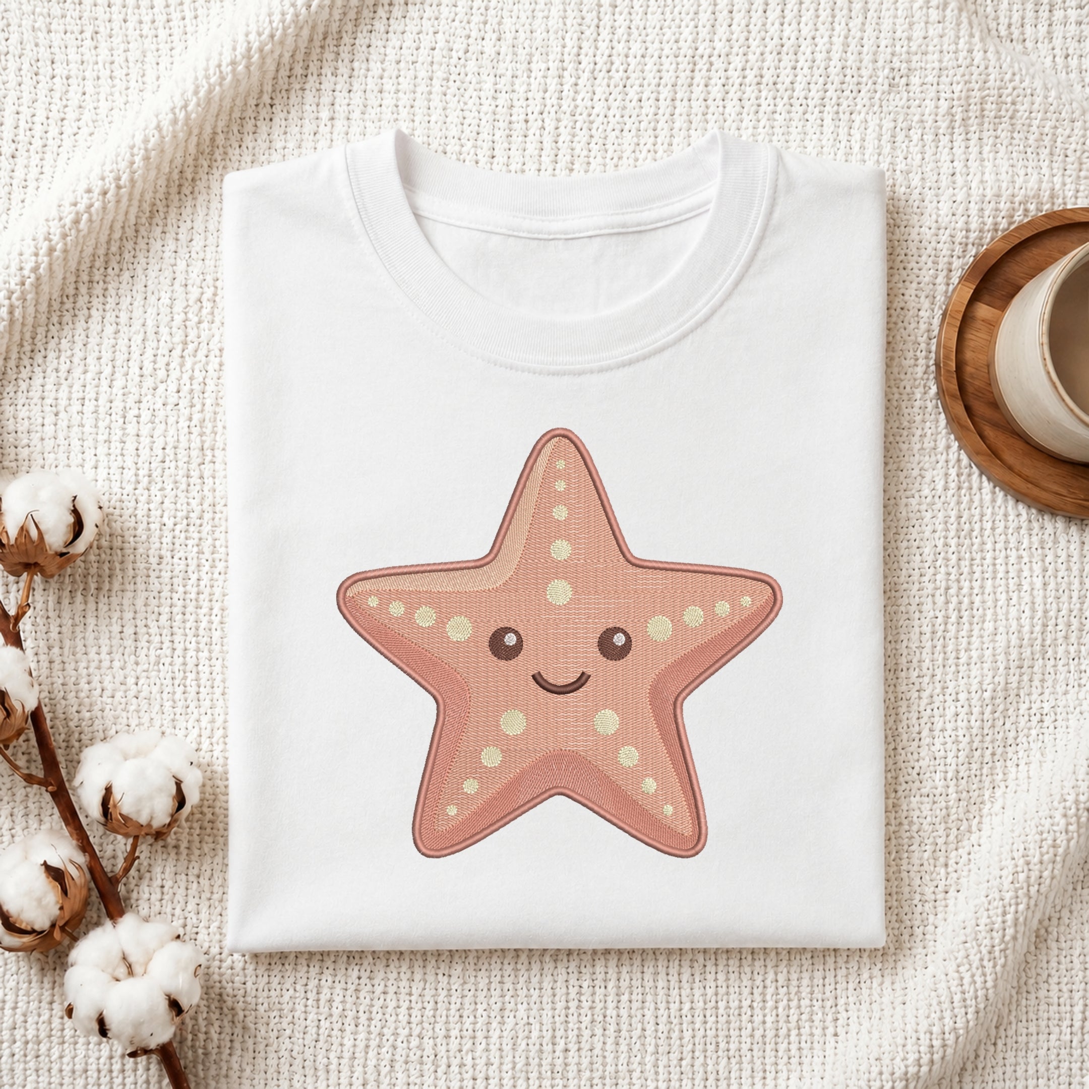 A folded white crewneck t-shirt featuring a cute pinkish-orange starfish graphic with a smiling face, styled on a textured cream knit blanket with cotton bolls and a wooden cup in the background.