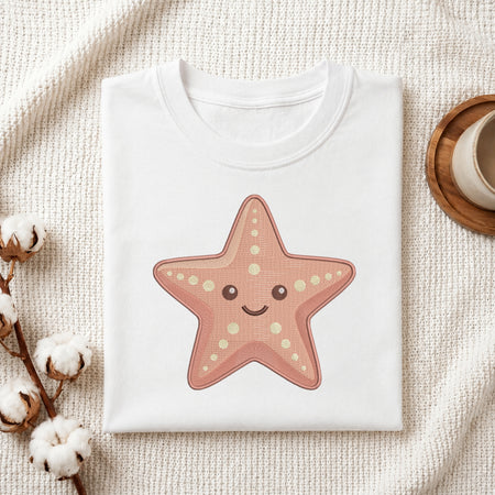 A folded white crewneck t-shirt featuring a cute pinkish-orange starfish graphic with a smiling face, styled on a textured cream knit blanket with cotton bolls and a wooden cup in the background.