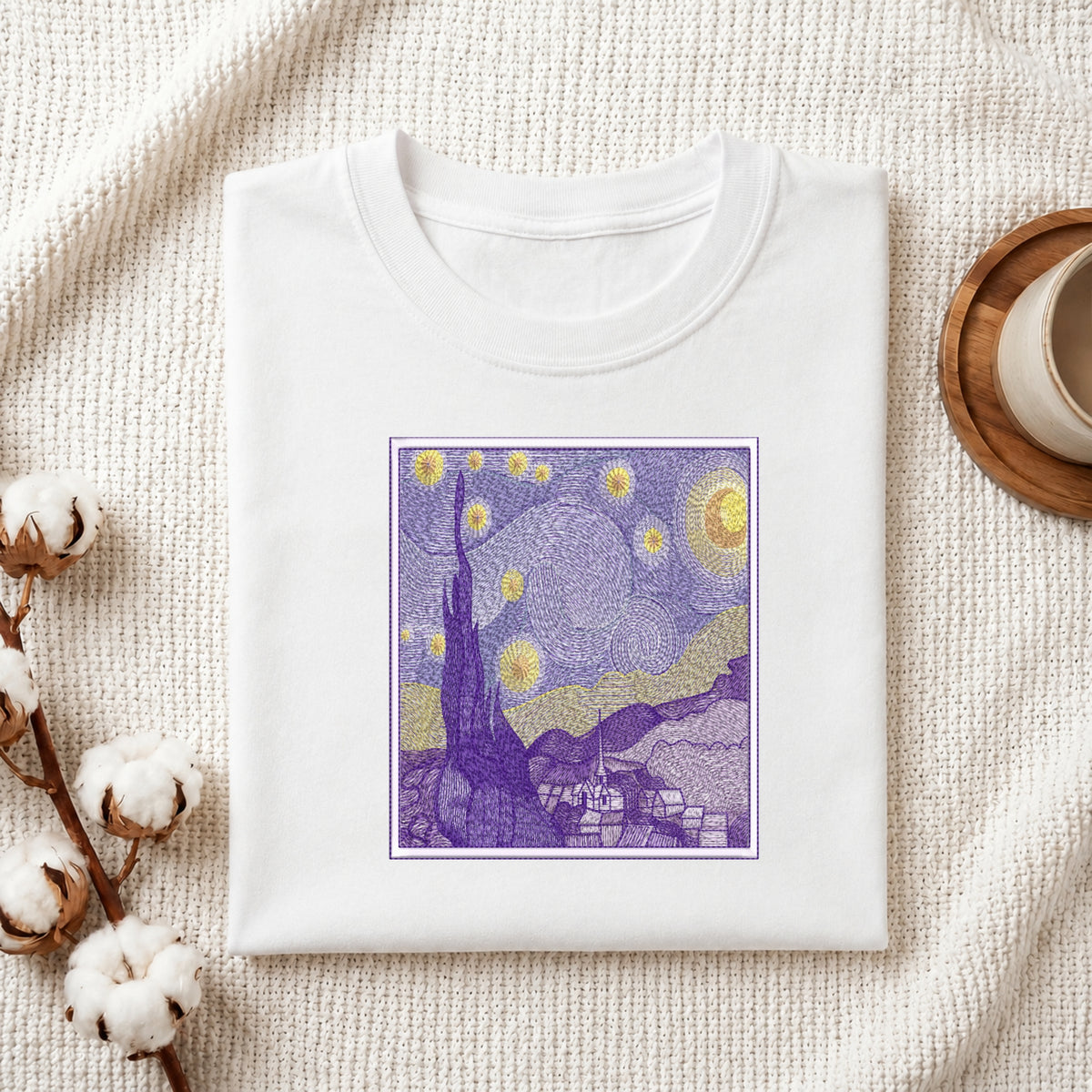 A folded white crewneck t-shirt displayed on a textured cream woven blanket, styled with cotton bolls on the left and a wooden cup on the right. The t-shirt features a square graphic print of a Van Gogh-inspired night scene in purple, yellow, and blue.