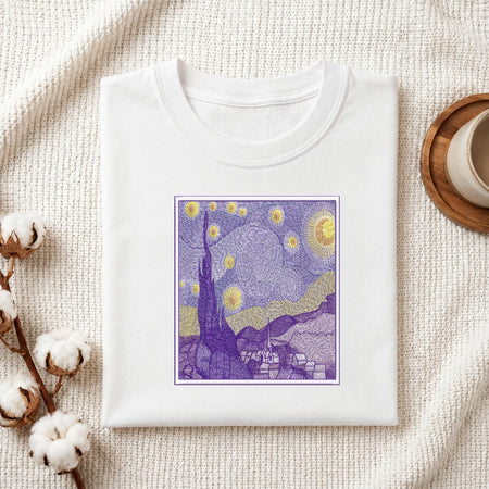 A folded white crewneck t-shirt displayed on a textured cream woven blanket, styled with cotton bolls on the left and a wooden cup on the right. The t-shirt features a square graphic print of a Van Gogh-inspired night scene in purple, yellow, and blue.