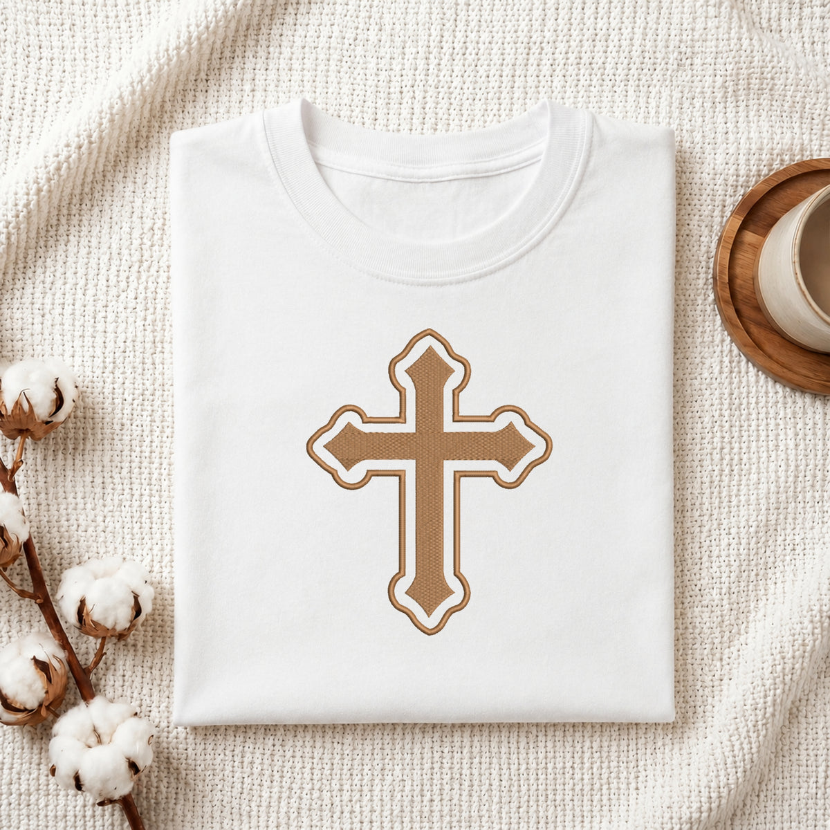 A neatly folded white crewneck t-shirt featuring the ornate brown and white Christian cross embroidery design centered on the front, styled on a textured knit blanket next to a cup and cotton stems.
