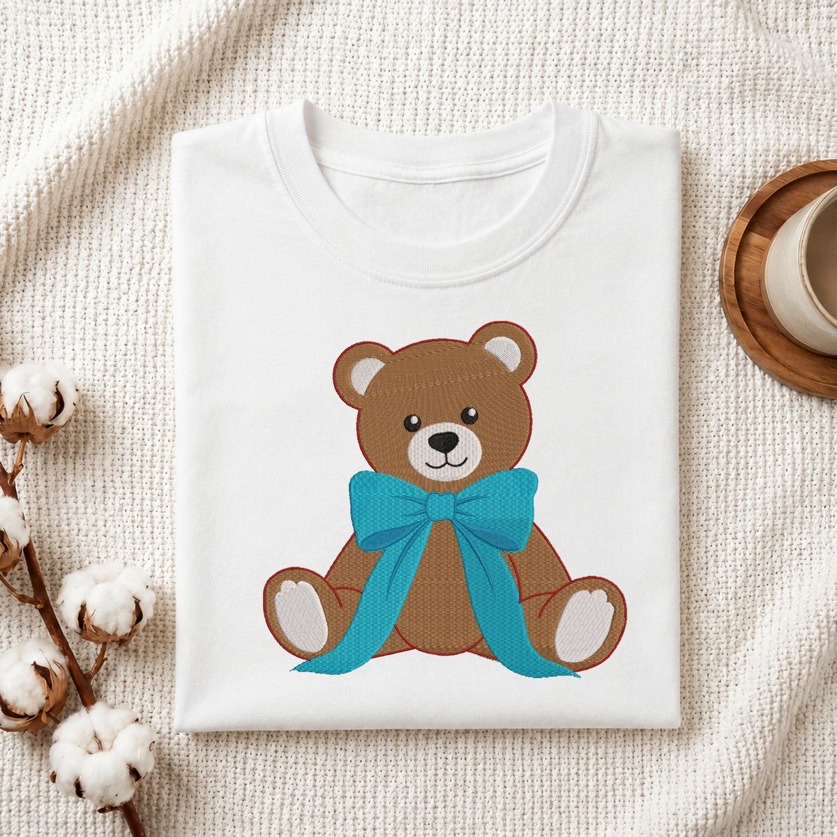 A neatly folded white crewneck t-shirt featuring a stitched brown teddy bear design wearing a large blue ribbon bow, styled with cotton branches and a wooden coaster.