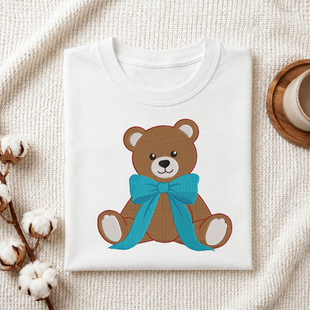 A neatly folded white crewneck t-shirt featuring a stitched brown teddy bear design wearing a large blue ribbon bow, styled with cotton branches and a wooden coaster.