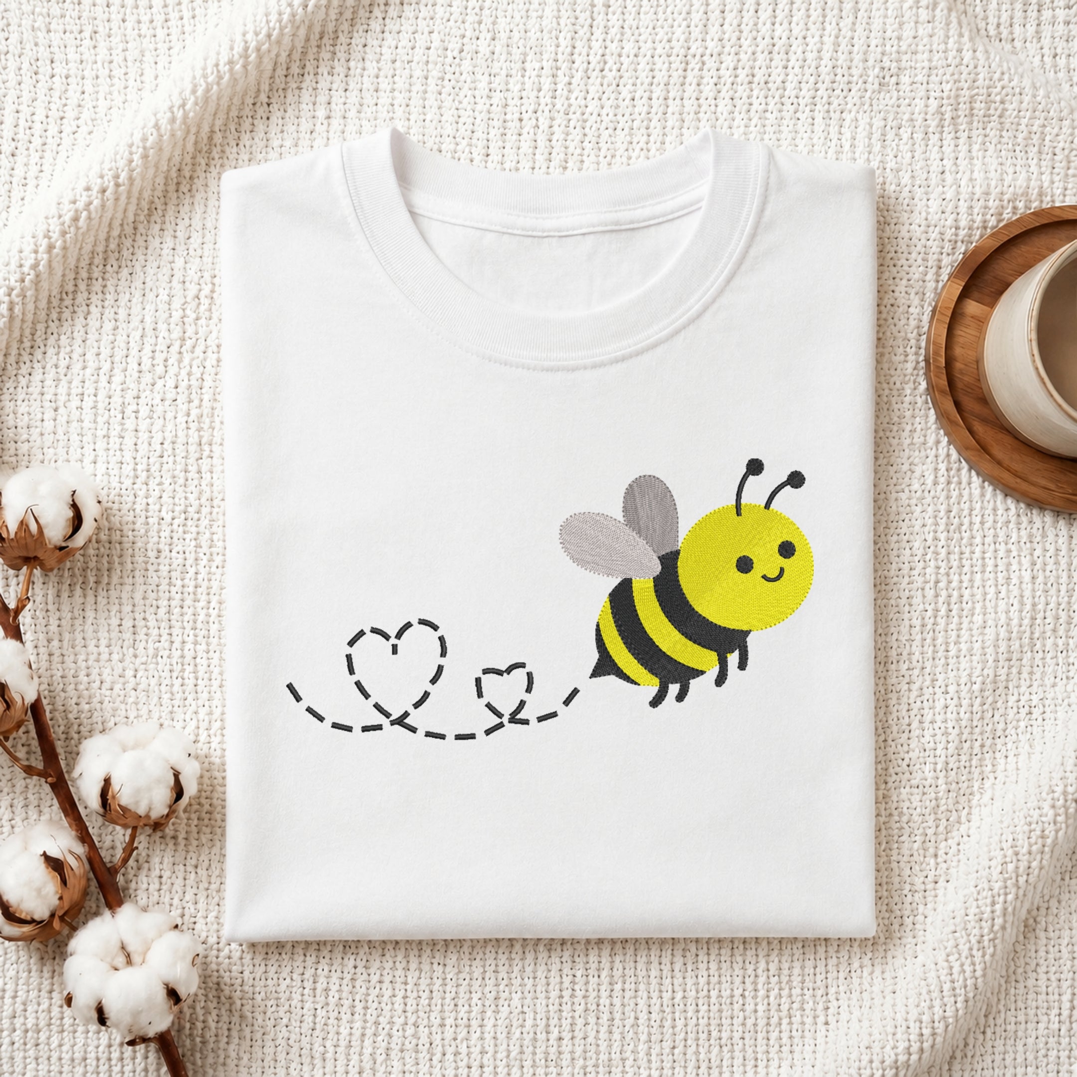 A folded white t-shirt with a cute cartoon bee graphic on the front, styled in a flat-lay on a textured cream fabric with cotton bolls and a wooden cup.