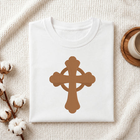 A neatly folded white crewneck t-shirt featuring the brown Celtic cross embroidery design centered on the front, styled on a textured knit blanket next to a cup and cotton stems.