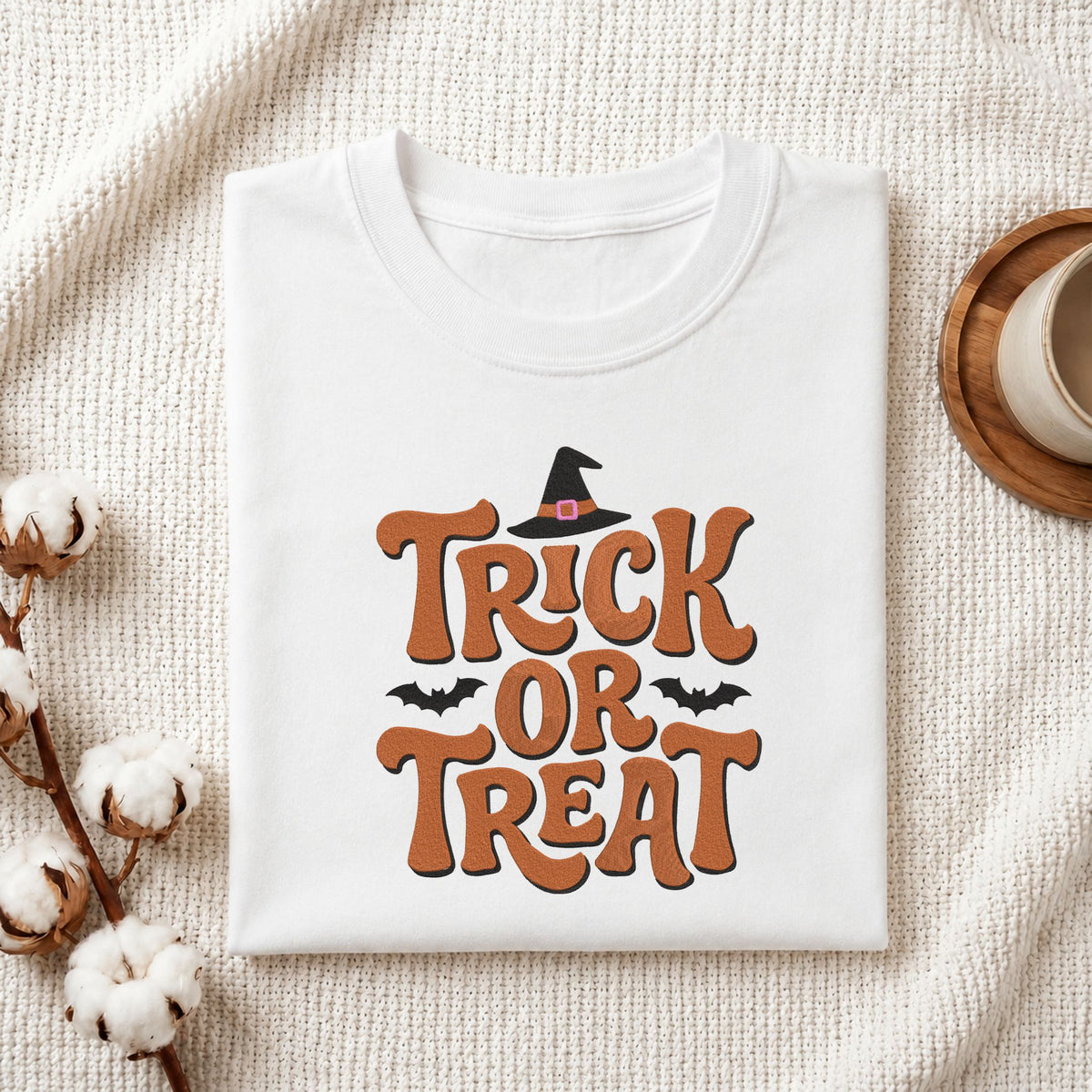 Folded white cotton t-shirt mockup featuring a "Trick or Treat" witch hat machine embroidery design, styled with cotton stems and a wooden cup