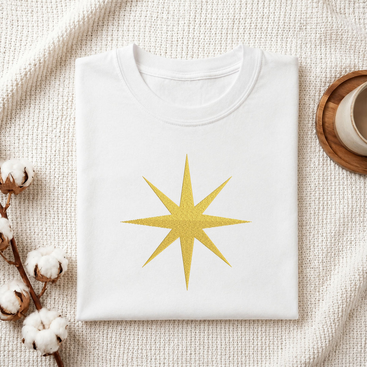  Folded white cotton t-shirt mockup featuring a golden swirling star machine embroidery design, styled with cotton stems and a wooden cup   