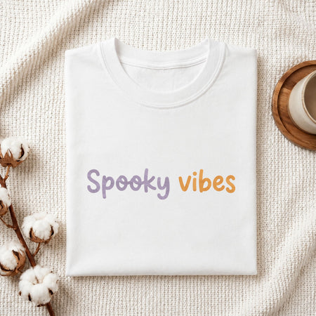 Folded white cotton t-shirt mockup featuring a spooky vibes machine embroidery design, styled with cotton stems and a wooden cup