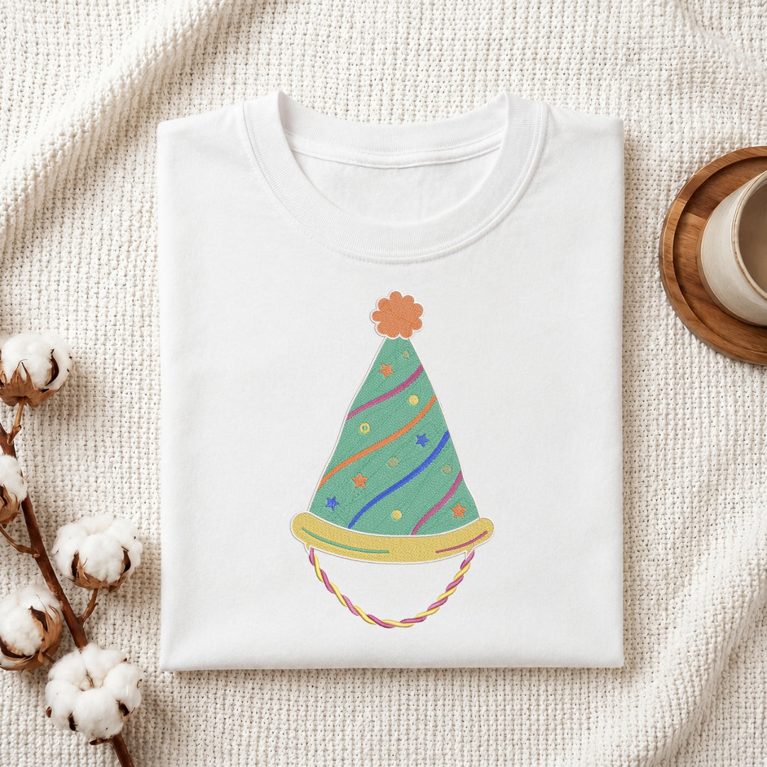 A folded white crewneck t-shirt with a colorful party hat graphic, styled on a textured cream knit blanket with cotton bolls and a wooden cup in the background.