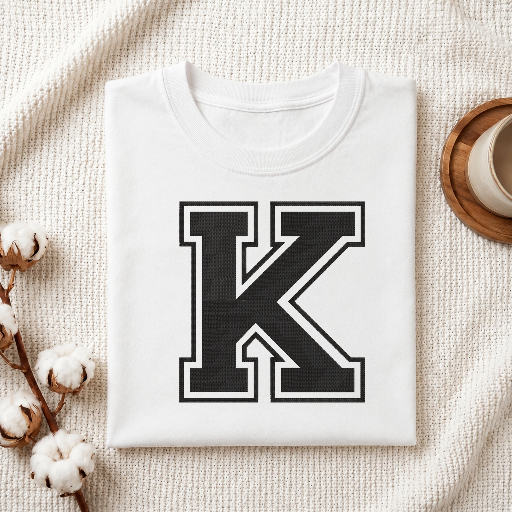 A folded white T-shirt with a large black block letter K outlined in white on the front, styled on a textured cream fabric with cotton bolls and a small cup at the side.