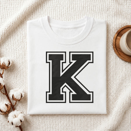 A folded white T-shirt with a large black block letter K outlined in white on the front, styled on a textured cream fabric with cotton bolls and a small cup at the side.