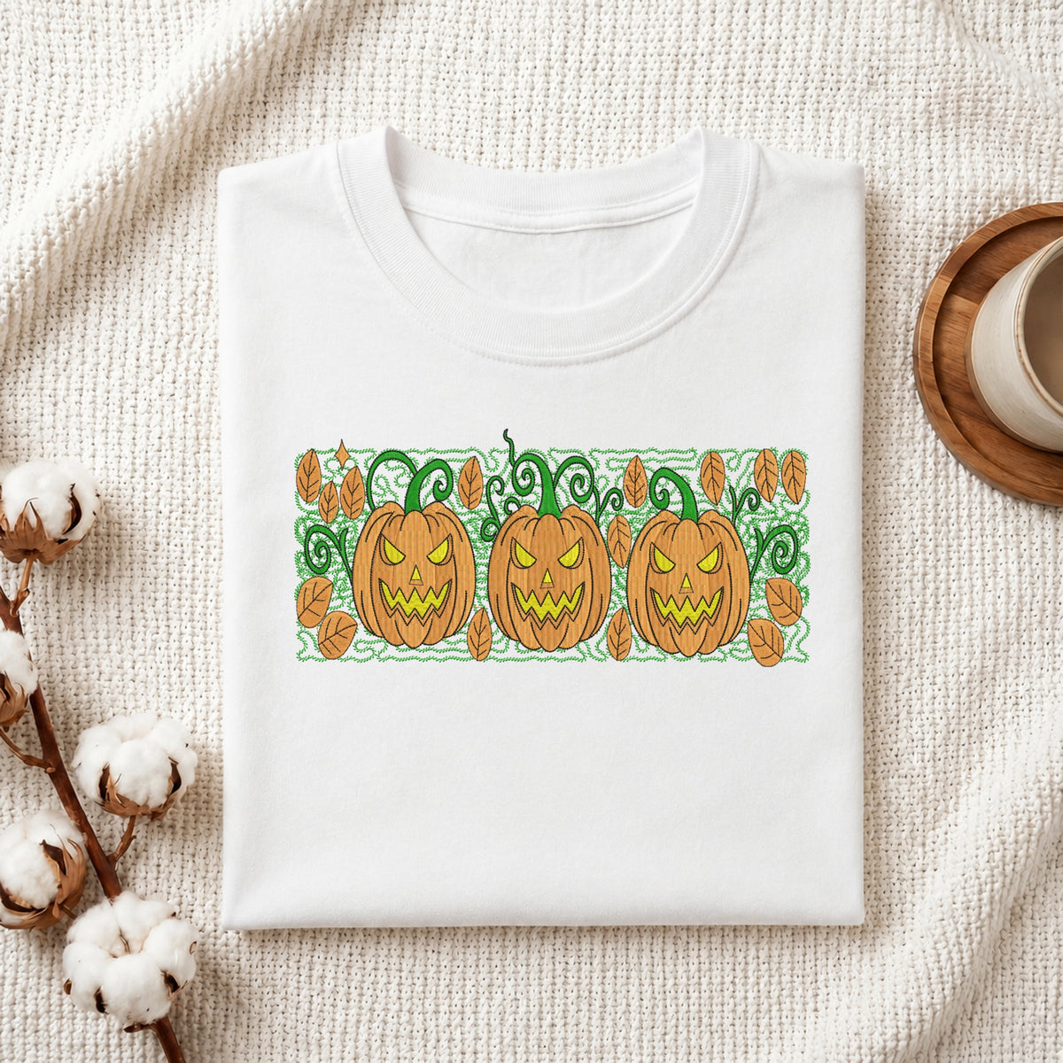 A folded white t-shirt with a Halloween graphic print of three carved pumpkins surrounded by autumn leaves and vines, styled on a textured cream fabric with cotton bolls and a mug.