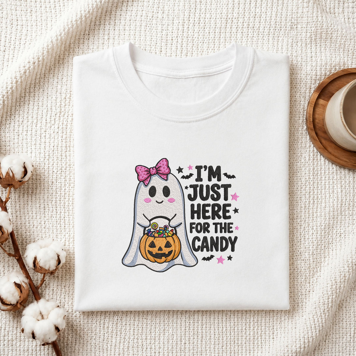 Folded white cotton t-shirt mockup featuring an "I'm Just Here for the Candy" cute ghost machine embroidery design, styled with cotton stems and a wooden cup   