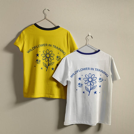 Two t-shirts hanging on a wall, one yellow and one white, both featuring the retro line art "Wildflower in Training" daisy graphic design.