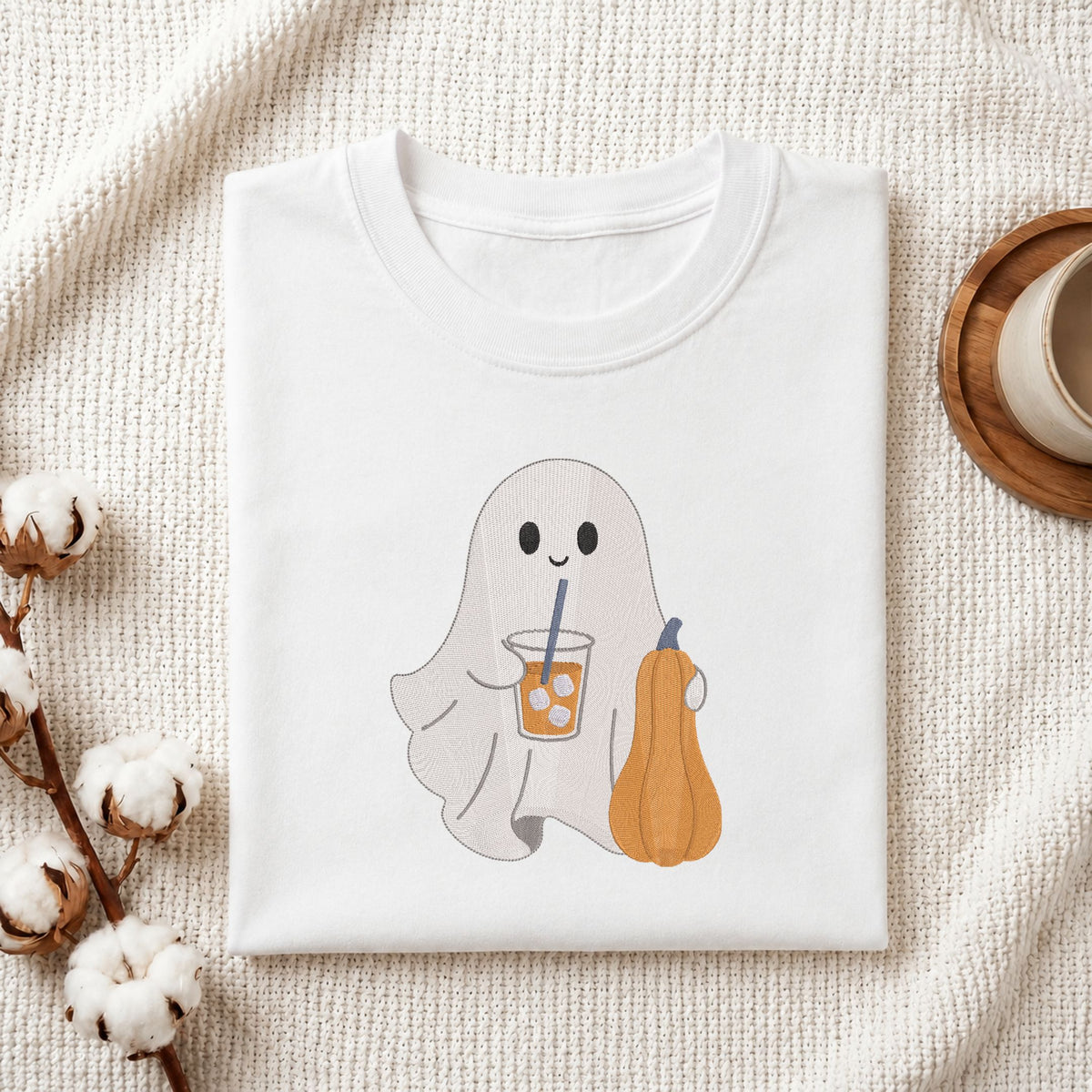 Folded white cotton t-shirt mockup featuring a cute ghost drinking iced coffee machine embroidery design, styled with cotton stems and a wooden cup.