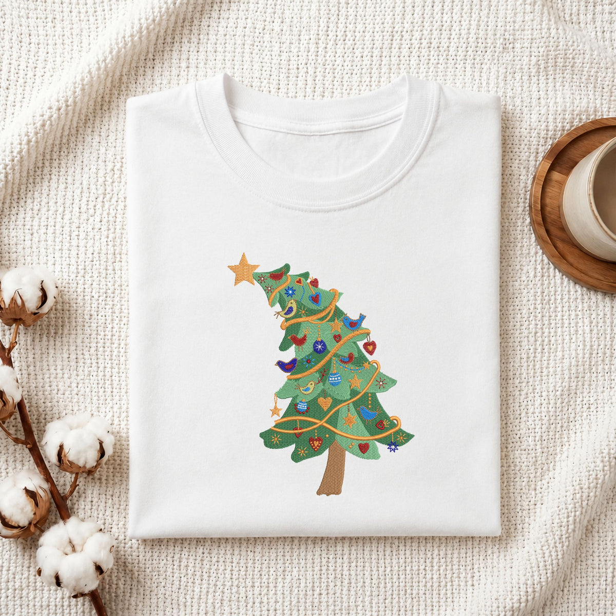 Folded white cotton t-shirt mockup featuring a tilted Christmas tree machine embroidery design, styled with cotton stems and a wooden cup.