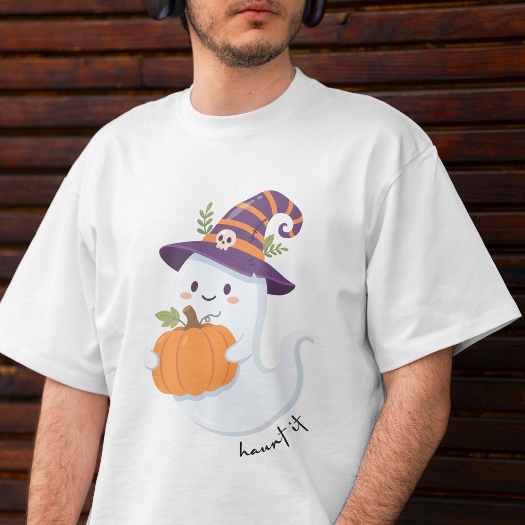 A man wearing a white t-shirt with a cute cartoon ghost wearing a purple witch hat and holding an orange pumpkin, with the text 'haunted it' printed below. The man is standing in front of a dark brown wooden slat wall.