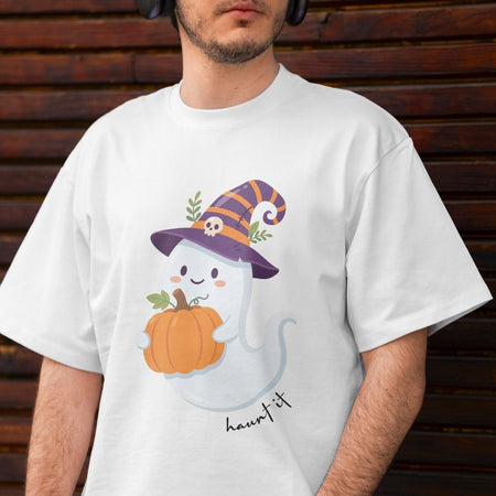 A man wearing a white t-shirt with a cute cartoon ghost wearing a purple witch hat and holding an orange pumpkin, with the text 'haunted it' printed below. The man is standing in front of a dark brown wooden slat wall.