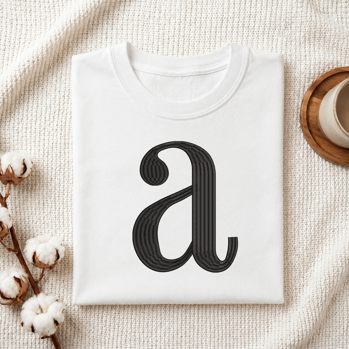 A folded white t-shirt with a black stylized lowercase 'a' graphic printed on the front, styled in a flat-lay on a textured cream knit blanket with cotton bolls and a wooden cup.