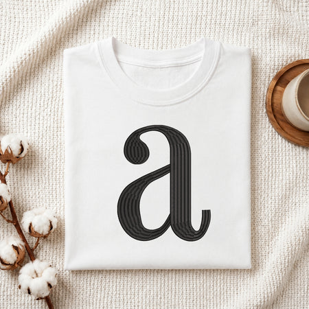 A folded white t-shirt with a black stylized lowercase 'a' graphic printed on the front, styled in a flat-lay on a textured cream knit blanket with cotton bolls and a wooden cup.