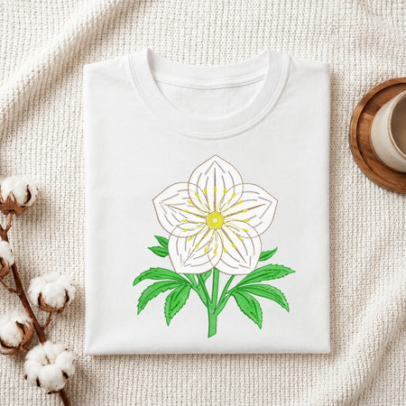 A folded white crewneck t-shirt featuring a hand-drawn style white flower with a yellow center and green leaves, displayed on a textured cream fabric background with decorative cotton bolls and a wooden cup.