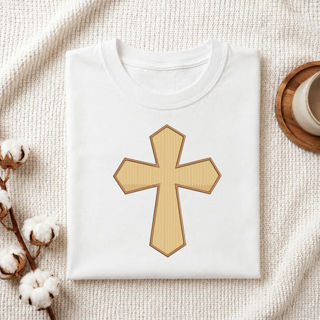 A neatly folded white crewneck t-shirt featuring the textured gold-yellow Christian cross embroidery design centered on the front, styled on a textured knit blanket next to a cup and cotton stems.