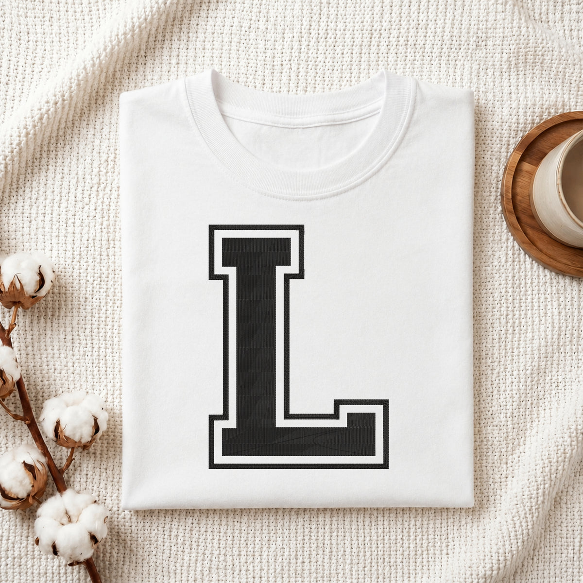 A folded white crewneck t-shirt with a large black block letter 'L' outlined in white on the front, styled on a textured cream knit blanket with cotton stems and a wooden cup in the background.
