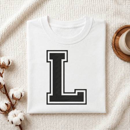 A folded white crewneck t-shirt with a large black block letter 'L' outlined in white on the front, styled on a textured cream knit blanket with cotton stems and a wooden cup in the background.