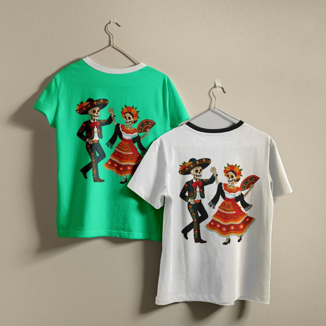 Two children's short-sleeve T-shirts hanging on hangers against a plain beige wall. The left shirt is bright green and shows a large Day of the Dead sugar skull couple graphic on the back.