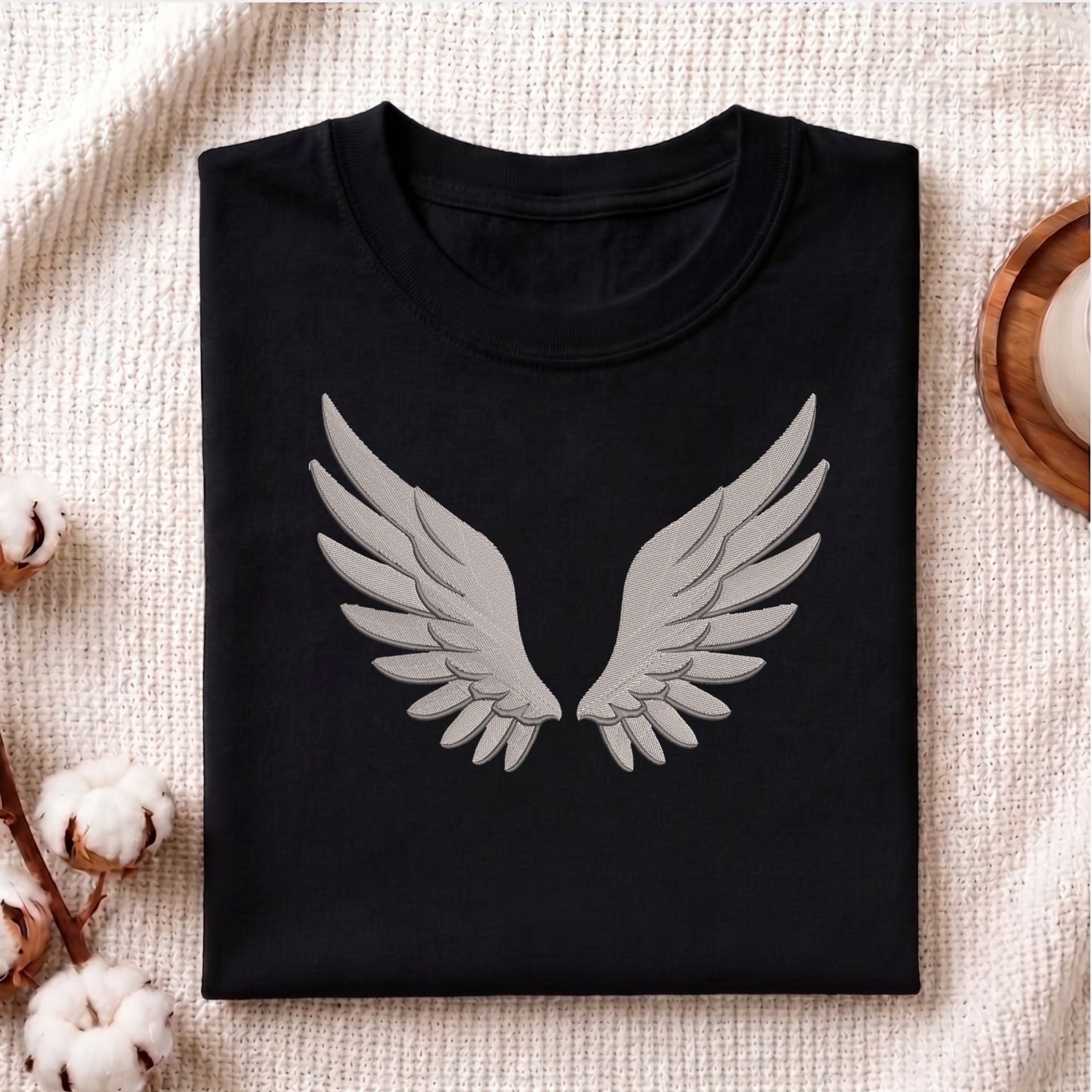 A folded black crewneck t-shirt with a large gray angel wings graphic on the front, displayed on a textured beige blanket with cotton bolls and a wooden tray as props.
