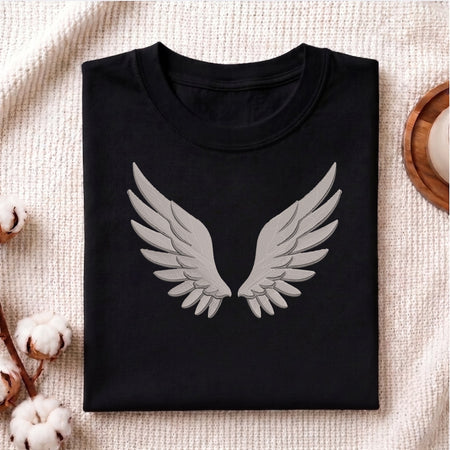 A folded black crewneck t-shirt with a large gray angel wings graphic on the front, displayed on a textured beige blanket with cotton bolls and a wooden tray as props.