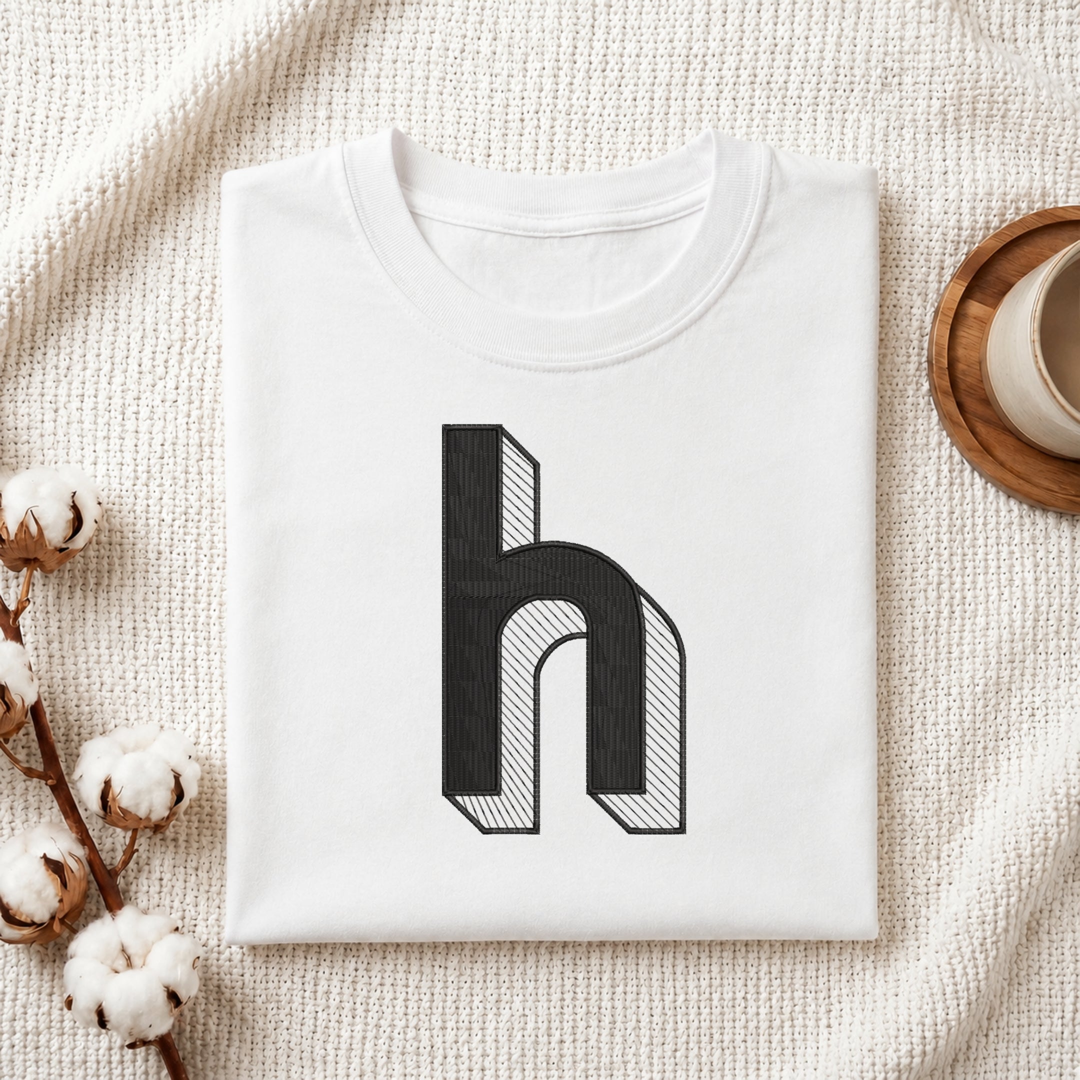A folded white t-shirt with a black 3D-effect letter 'h' graphic on the front, styled on a textured cream blanket with cotton bolls and a cup of coffee.