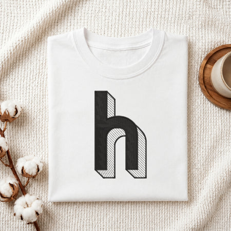 A folded white t-shirt with a black 3D-effect letter 'h' graphic on the front, styled on a textured cream blanket with cotton bolls and a cup of coffee.