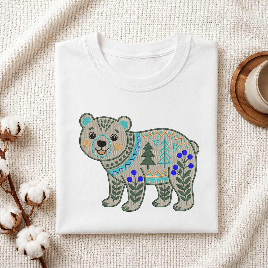A folded white crewneck t-shirt featuring a colorful geometric patterned bear graphic, styled on a textured cream fabric with cotton bolls and a wooden cup.