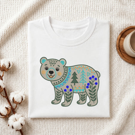 A folded white crewneck t-shirt featuring a colorful geometric patterned bear graphic, styled on a textured cream fabric with cotton bolls and a wooden cup.