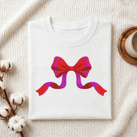 A folded white crewneck t-shirt with a red and purple ribbon graphic on the chest, styled on a textured cream knit blanket with cotton bolls and a wooden cup in the background.