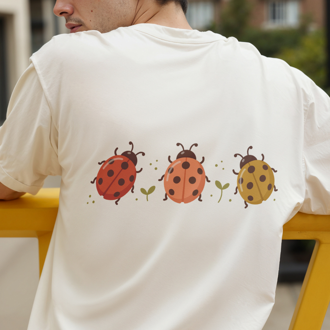 A man wearing a cream-colored t-shirt, viewed from behind, featuring a graphic print of three ladybugs in red, orange, and yellow with green leaves on the back.