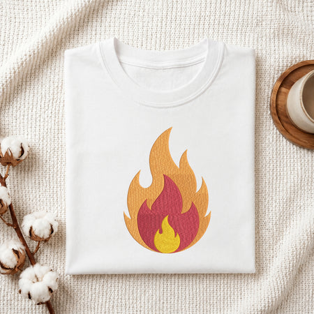 A folded white T-shirt with a stylized flame graphic in orange, yellow, and red on the front, displayed on a textured cream fabric with cotton stems and a small wooden cup at the edges.
