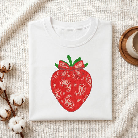 A folded white crewneck t-shirt featuring a red strawberry graphic with a paisley bandana pattern and green leaves, styled on a textured cream blanket with cotton bolls and a wooden cup.