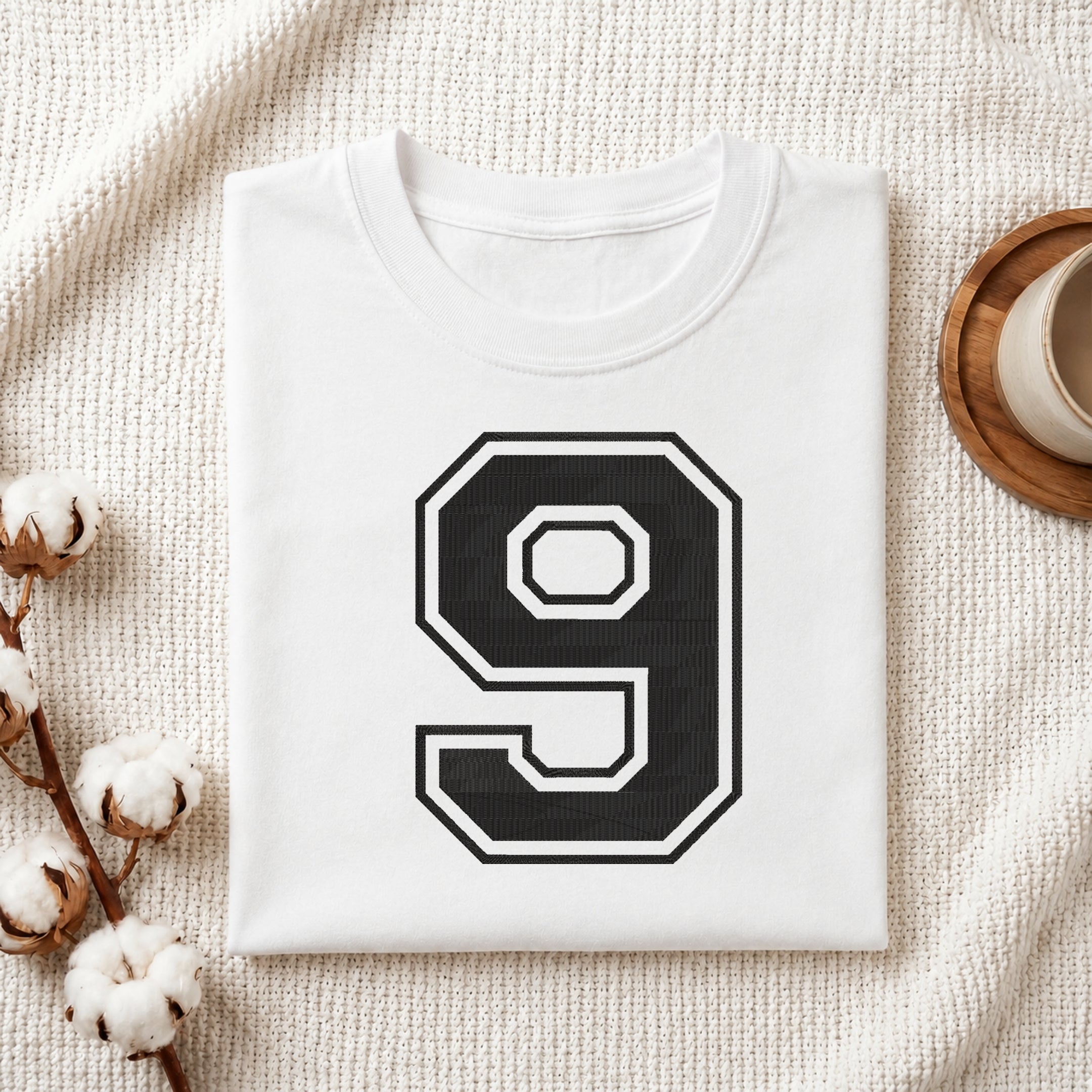 A folded white T-shirt with a large black and white number 9 printed on the front, styled on a textured cream fabric with cotton stems and a small cup in the background.