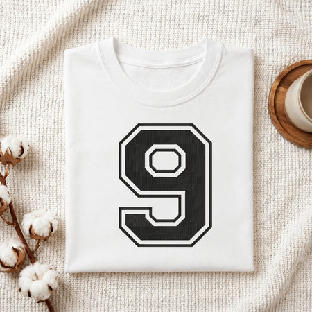 A folded white T-shirt with a large black and white number 9 printed on the front, styled on a textured cream fabric with cotton stems and a small cup in the background.